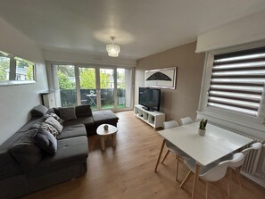A modern living room with a grey sectional sofa, a small round table, a TV on a white stand, and a dining area with a white table and chairs. - Vrbo Property (Lingolsheim)