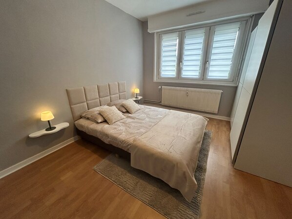 A bedroom with a bed, bedside lamps, a window with blinds, and a radiator. - Vrbo Property (Lingolsheim)