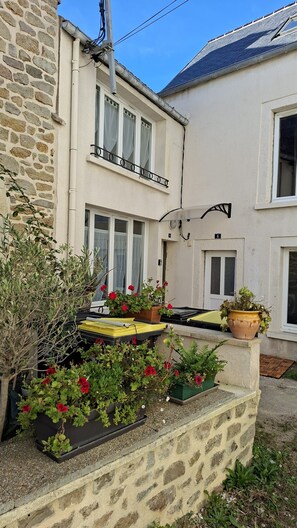 A two-story building with a stone facade, a balcony with a metal railing, and potted plants with red flowers. - ⚓ Charming fisherman's cottage in the heart of Saint-Vaast-la-Hougue (Saint-Vaast-la-Hougue)