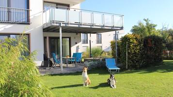 Ein modernes, zweistöckiges Gebäude mit überdachter Terrasse, Gartenmöbeln und einem gepflegten Rasen.
