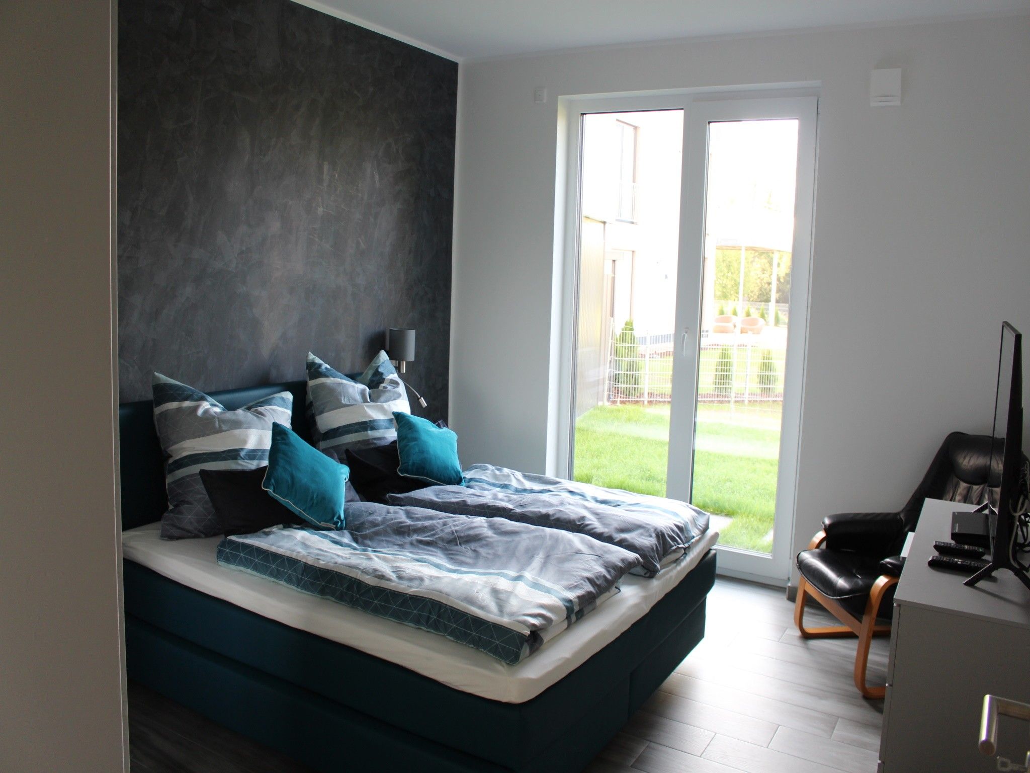 Une chambre moderne avec un grand lit, un bureau avec un ordinateur et une vue sur une pelouse verdoyante, visible à travers une porte coulissante.