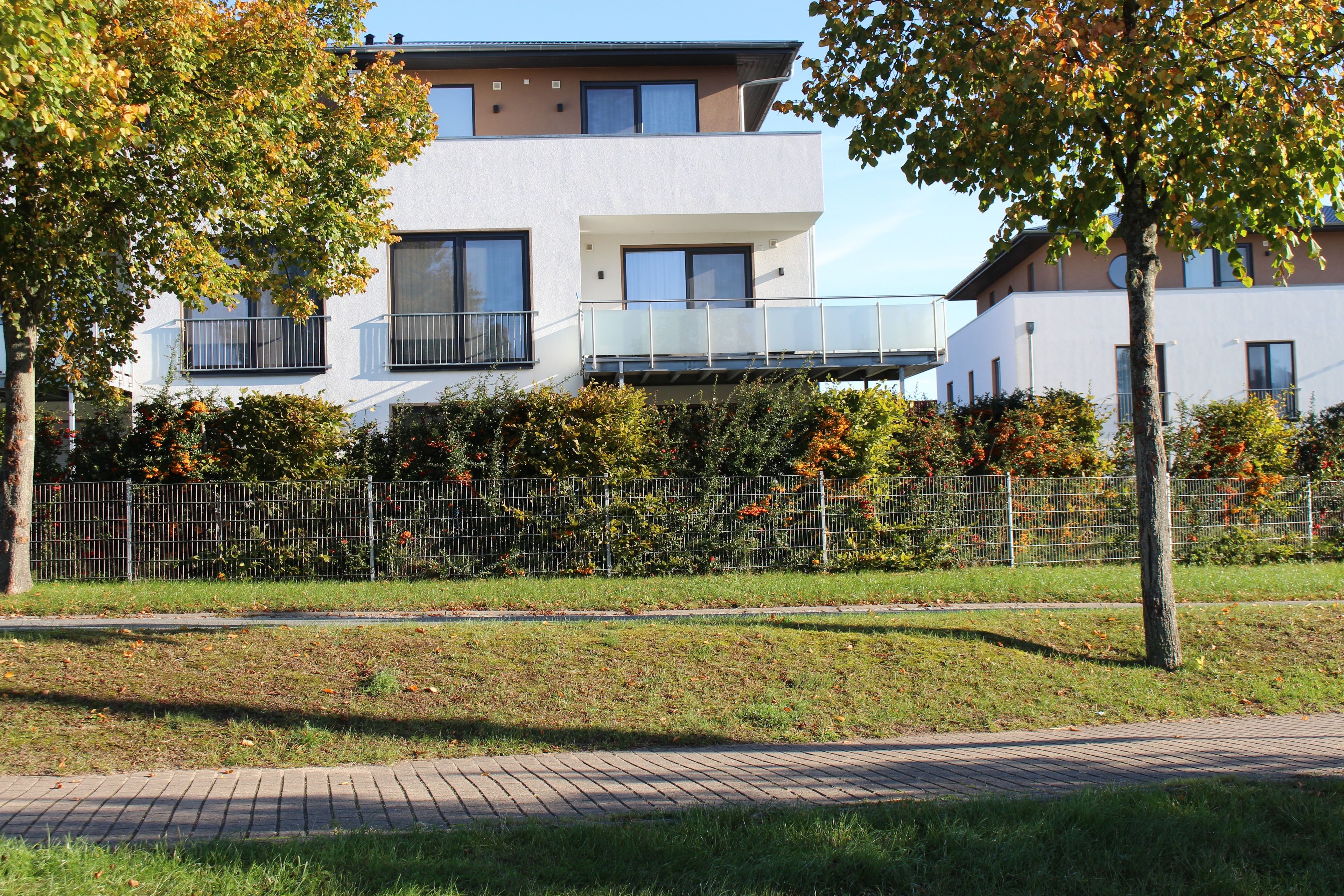 Ein modernes, zweistöckiges Wohnhaus mit Balkon, umgeben von einem Metallzaun und Bäumen.