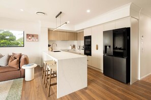 A modern kitchen with a central island, built-in appliances, and a dining area with a sofa and chairs. - Marea by Khove - Kingscliff (Kingscliff)