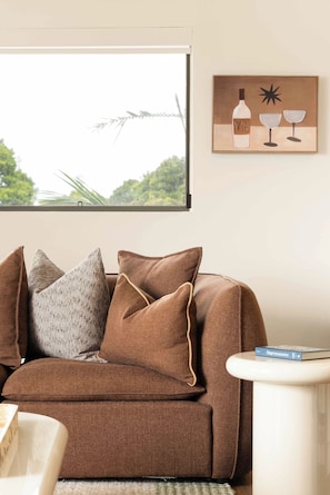 A brown sofa with multiple cushions, a framed artwork above it, and a book on a side table. - Marea by Khove - Kingscliff (Kingscliff)