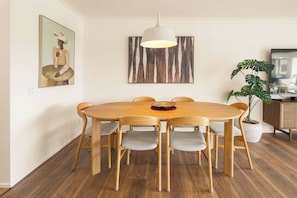 A dining area with a round wooden table, six chairs, a hanging light fixture, and artwork on the walls. - Marea by Khove - Kingscliff (Kingscliff)
