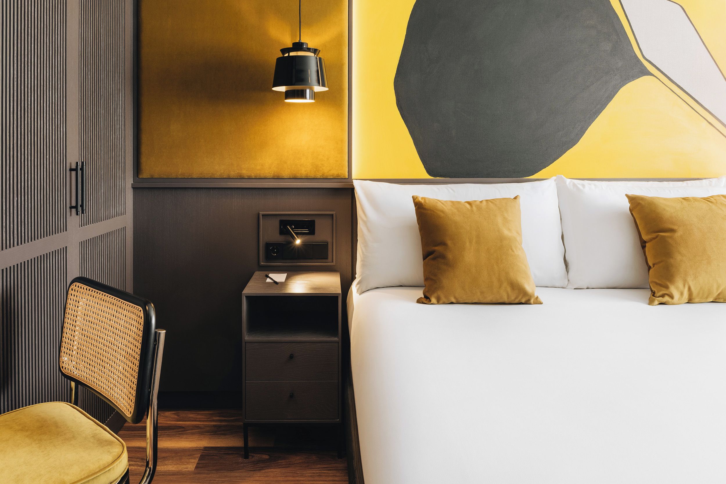 A hotel room with a bed, bedside table, chair, and a wall-mounted lamp.