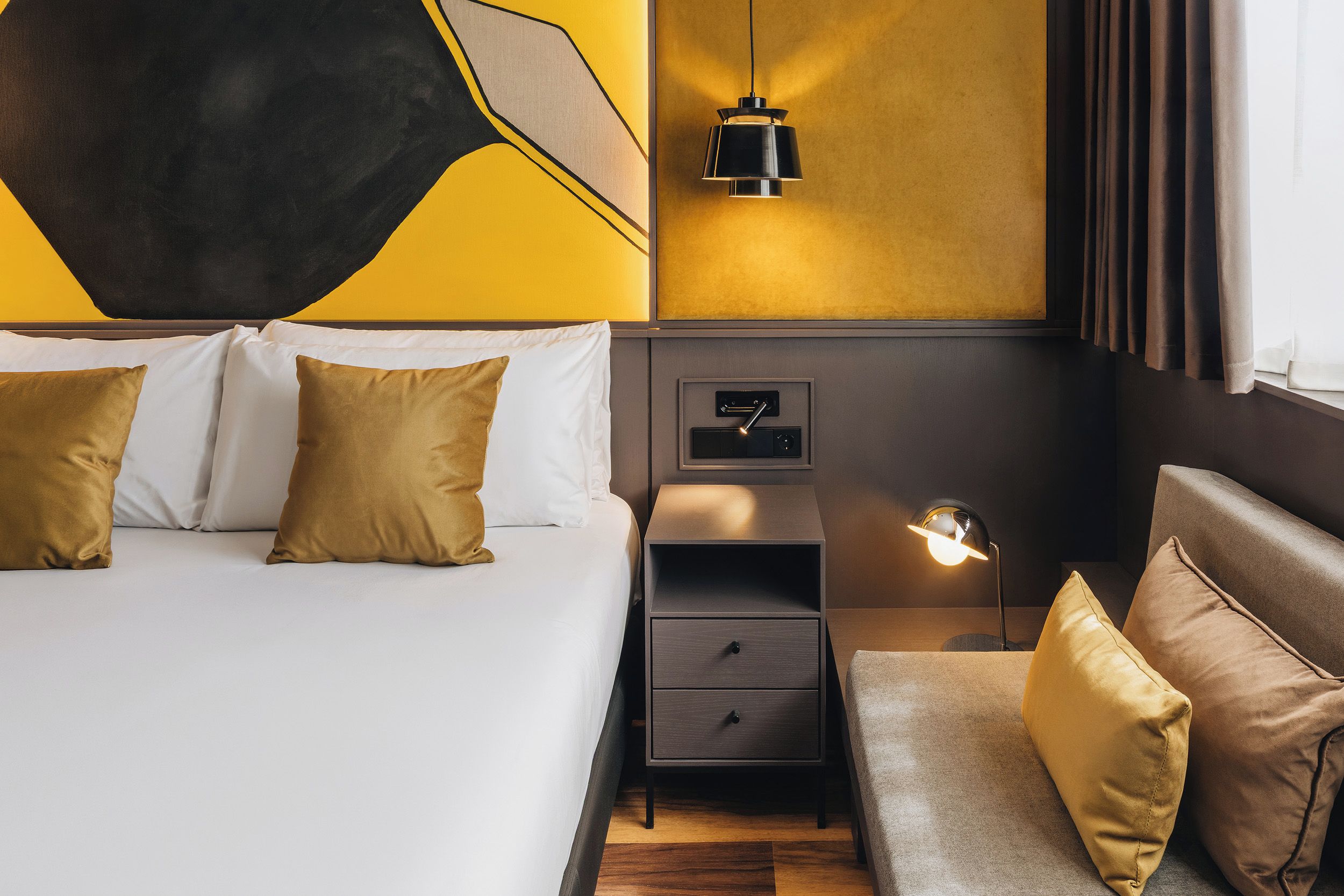 A hotel room with a bed, bedside table, lamp, and a wall-mounted control panel.