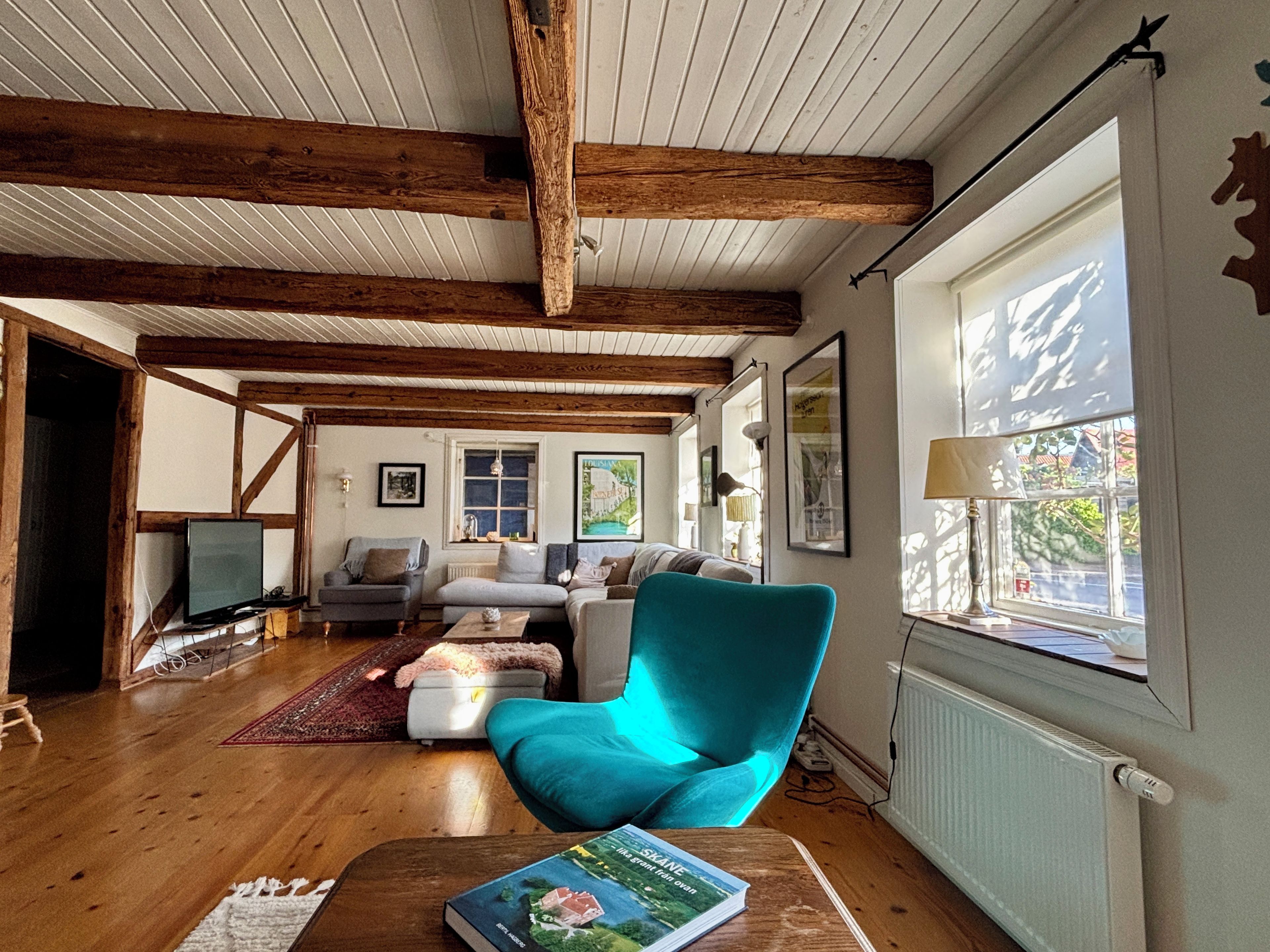 A cozy living room with wooden beams, a teal armchair, a fireplace, and a large window with a view.