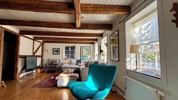 A cozy living room with wooden beams, a teal armchair, a fireplace, and a large window with a view.