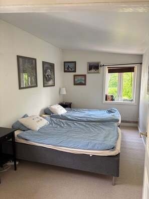 A bedroom with two beds, a nightstand, a lamp, and a window with a view of greenery.