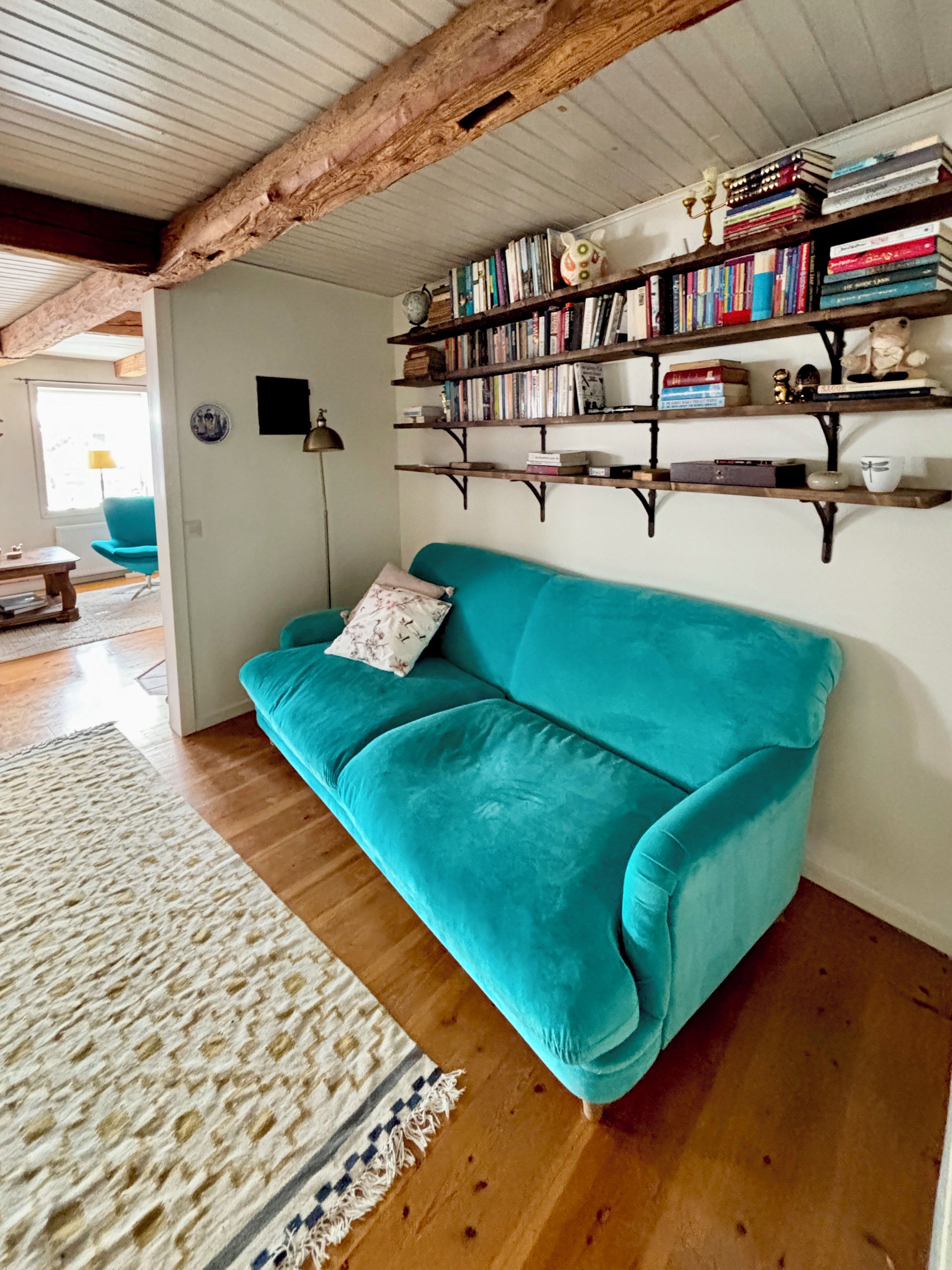 A teal sofa in a room with wooden floors, a patterned rug, and a bookshelf with various items.