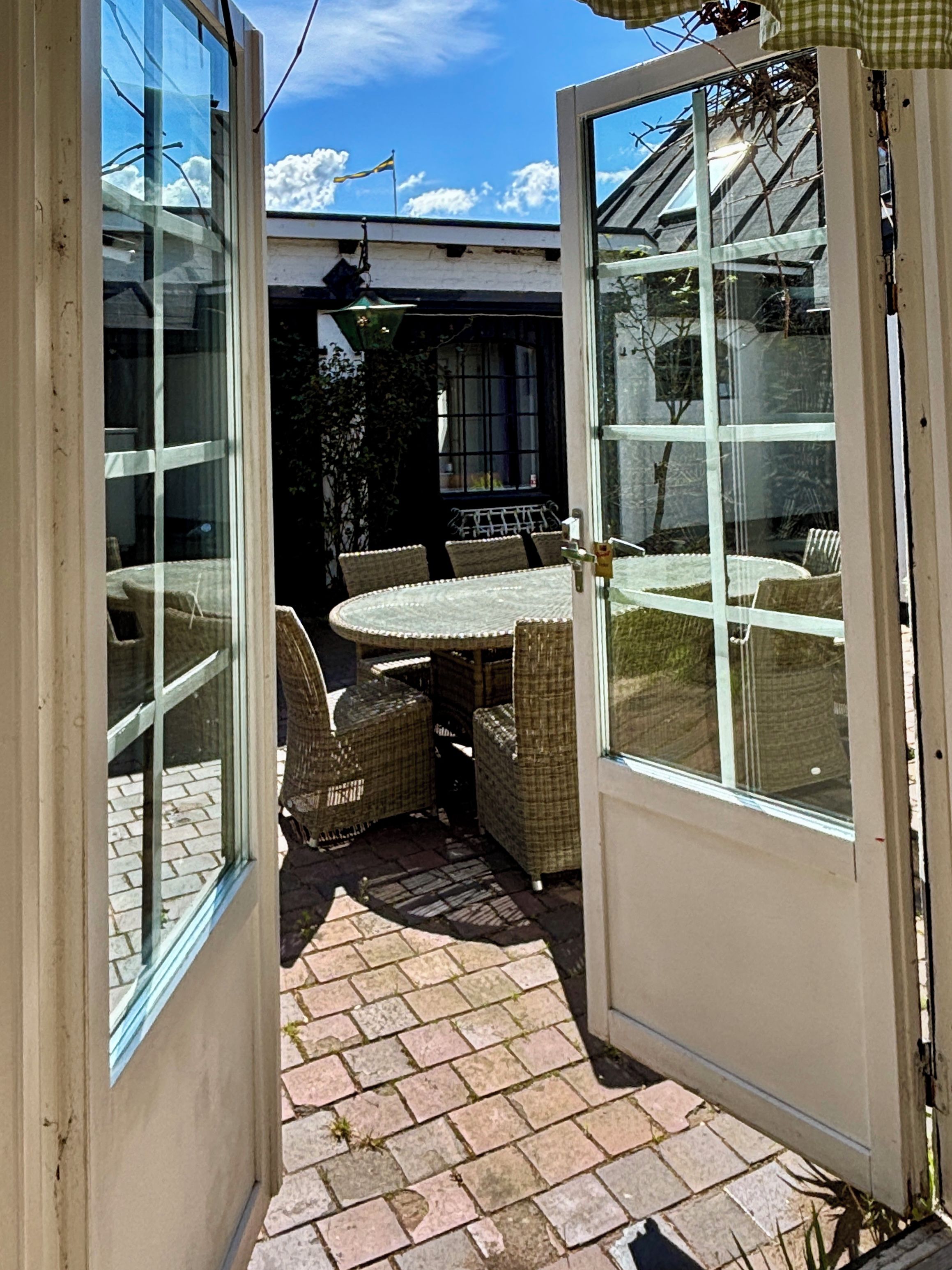 En patio med et bord og stole, en belagt sti i mursten og en glasdør, der fører til et udendørs område.