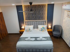 A modern hotel room with a large bed, a grey upholstered bench, a grey armchair, and a wall-mounted air conditioning unit. - Majestic Inn (Guwahati)