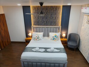 A modern hotel room with a large bed, two bedside tables with lamps, a grey upholstered armchair, and a ceiling fan. - Majestic Inn (Guwahati)
