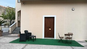 Un bâtiment à la façade beige, une porte en bois, un petit espace extérieur aménagé avec deux chaises et une table, et une pelouse artificielle verte.