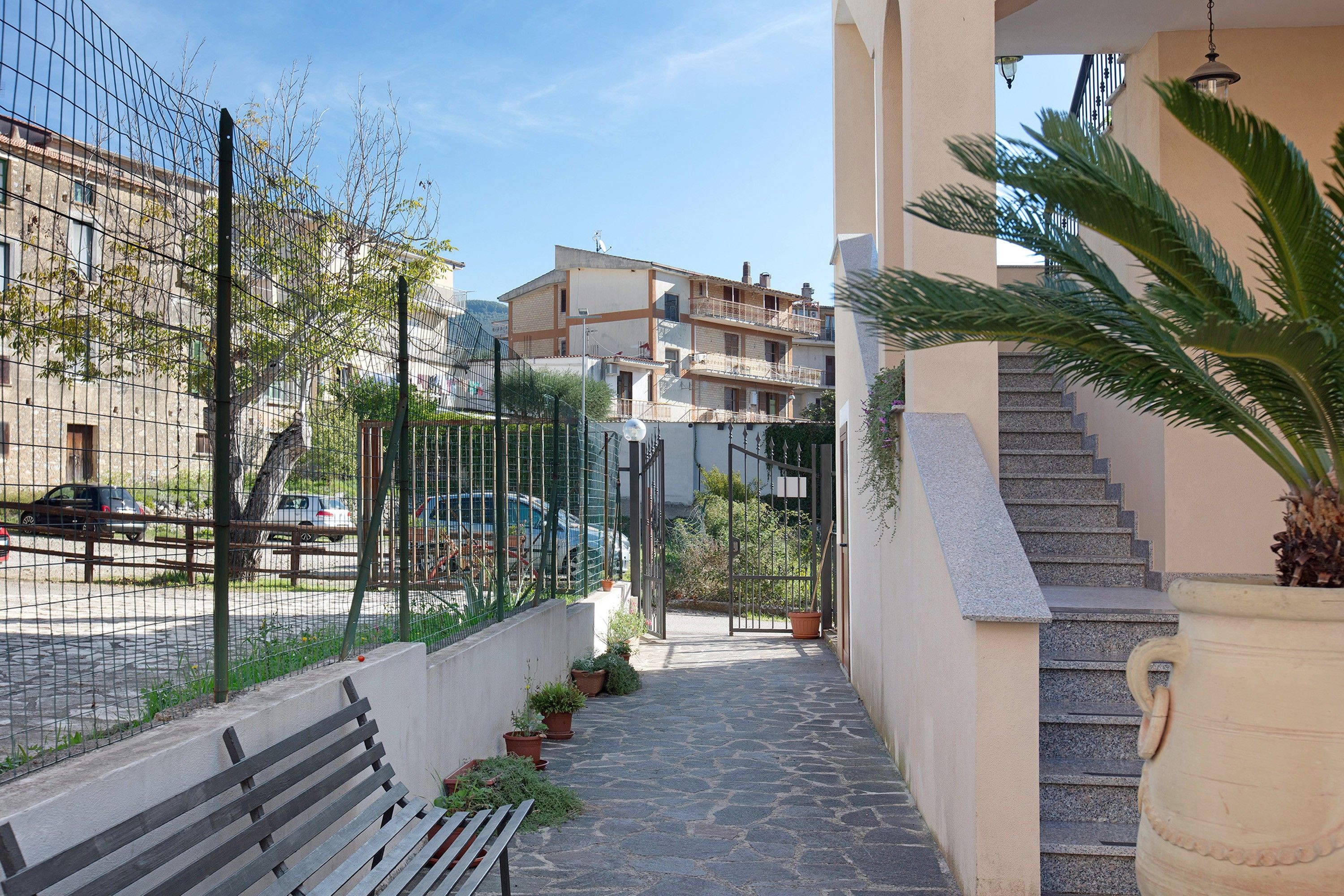Un étroit sentier mène à une entrée fermée par un portail, de part et d’autre se trouvent un banc et des plantes en pot, avec un escalier sur la droite et un bâtiment doté d’un balcon sur la gauche.
