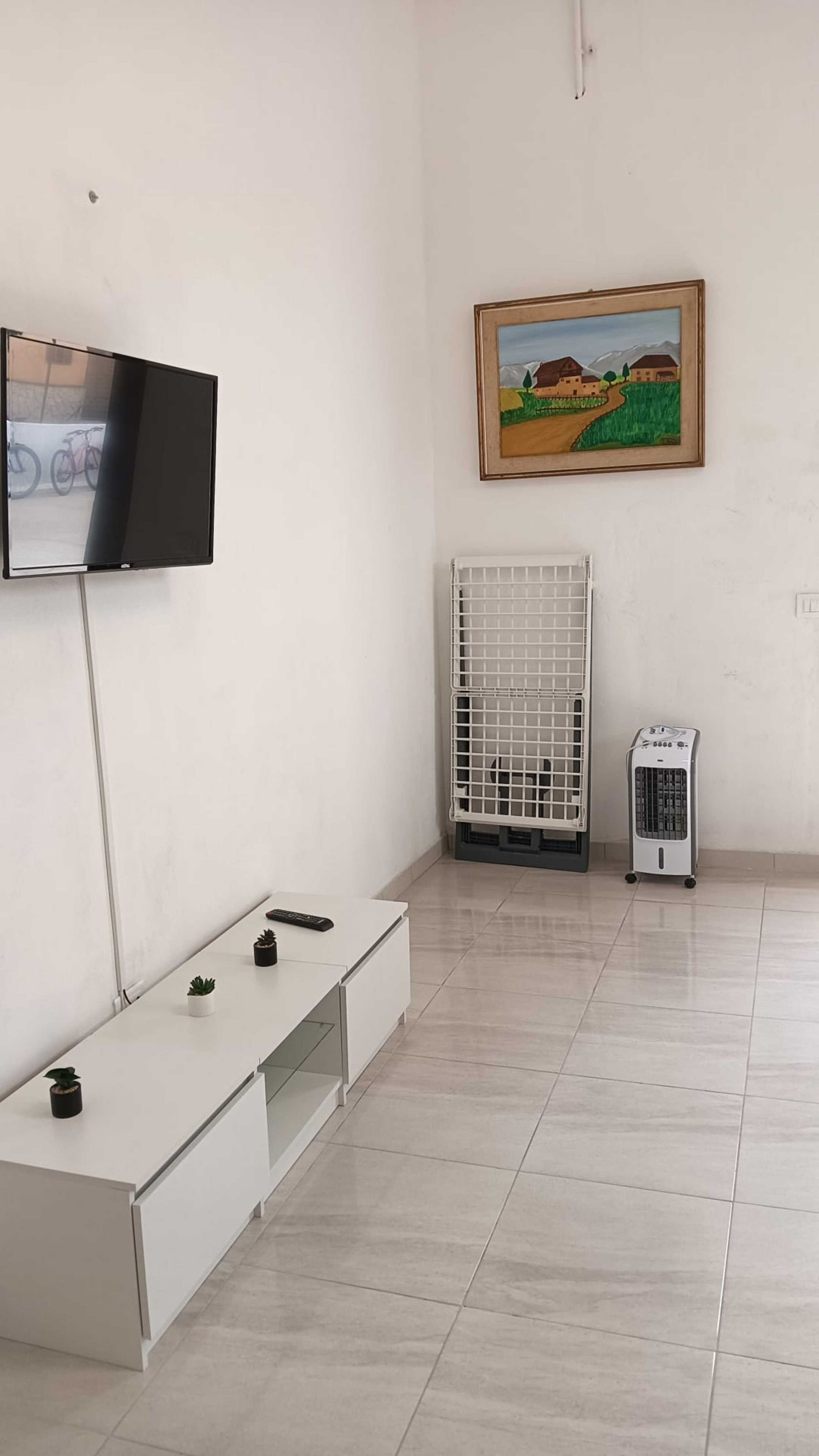 Une pièce dotée d’un téléviseur à écran plat fixé au mur, d’une armoire blanche avec des tiroirs, d’un radiateur, d’un chauffage d’appoint et d’un tableau encadré.