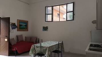 Un espace de vie compact comprenant un lit, une table à manger, un coin cuisine et une fenêtre donnant sur des immeubles.