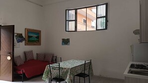 Un espace de vie compact comprenant un lit, une table à manger, un coin cuisine et une fenêtre donnant sur des immeubles.