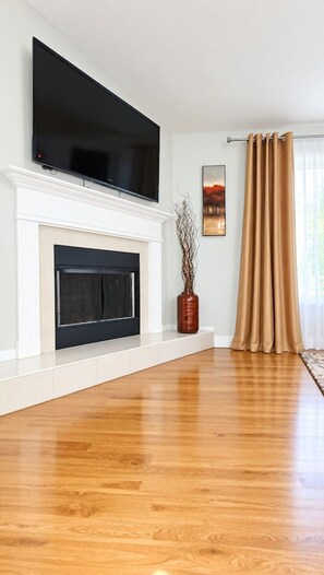 Un salon avec un téléviseur à écran plat fixé au-dessus d’une cheminée, un vase décoré de branches et une fenêtre avec des rideaux.
