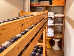 Un lit superposé avec un matelas à carreaux, une tête de lit en bois et une étagère où sont pliées des serviettes et des oreillers.