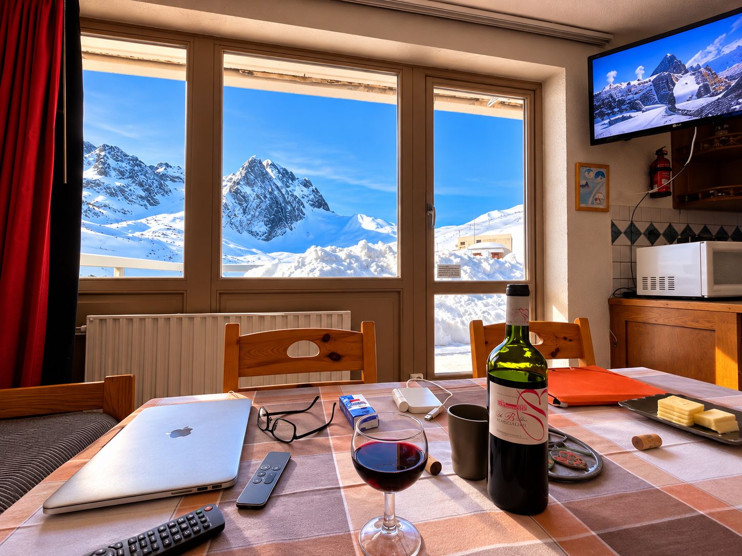 Un comedor con una mesa preparada para dos personas, una laptop, una copa de vino, controles remotos y una botella de vino.