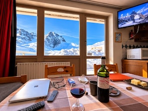 Un comedor con una mesa preparada para dos personas, una laptop, una copa de vino, controles remotos y una botella de vino.