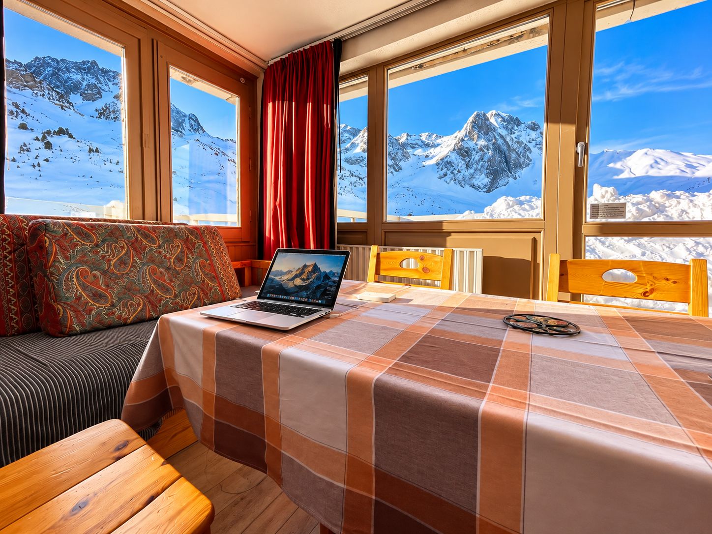 Una habitación con cama, una laptop sobre la cama, una ventana con vista a la montaña y una cortina roja.