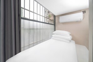 A hotel room with a bed, a window with blinds, an air conditioning unit, and a view of buildings outside.