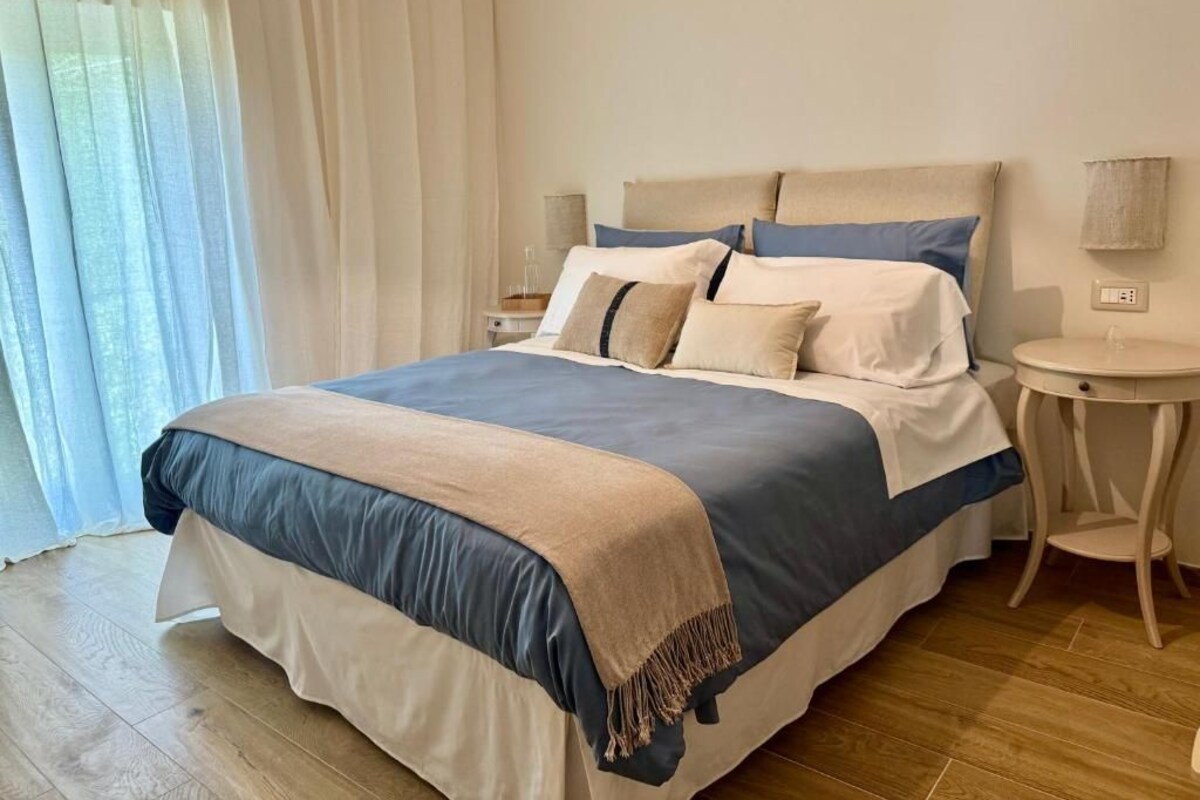 Un lit bien fait, recouvert d’une couvre-lit bleue et beige, une table de chevet avec une lampe, et une fenêtre ornée de rideaux légers.