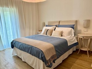Un lit bien fait, recouvert d’une couvre-lit bleue et beige, une table de chevet avec une lampe, et une fenêtre ornée de rideaux légers.