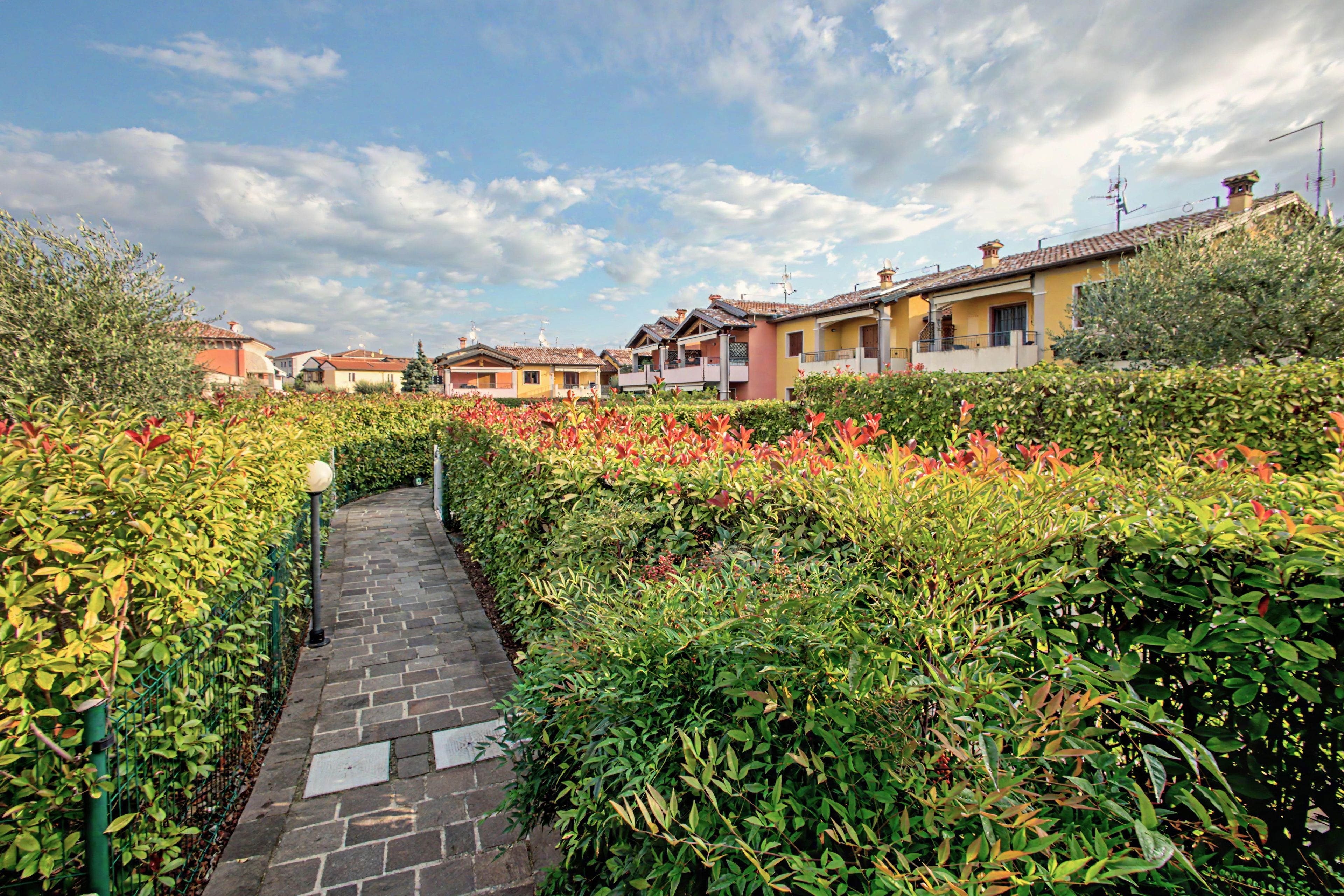 Un sentier traverse un jardin où les haies sont soigneusement taillées, et longe une rangée de bâtiments traditionnels aux toits de tuiles.