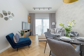 A modern living room with a blue sofa, grey armchairs, a dining table set for four, and a TV mounted on the wall. - Mrzezyno | Nadmorska 7F | Comfortable Apartment | Air conditioning | Parking (Mrzeżyno)