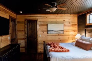 Ein Zimmer in einer Holzhütte mit einem Bett, einem Deckenventilator, einem Gemälde und einem Fernseher.