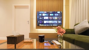 A modern living room with a flat-screen TV mounted on a wooden wall, a green sofa, a glass coffee table with remote controls, a vase with flowers, and a woven basket. - Luxury 1BR  Self Entry Near
KAFD Riyadh Blvd (Riyadh)