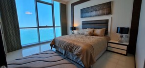 A bedroom with a large window, a bed with a headboard, bedside tables, and a wall-mounted artwork. - Amazing Full Sea View High floor apartment 3bedrooms 4bathrooms.5star facilities (Manama)