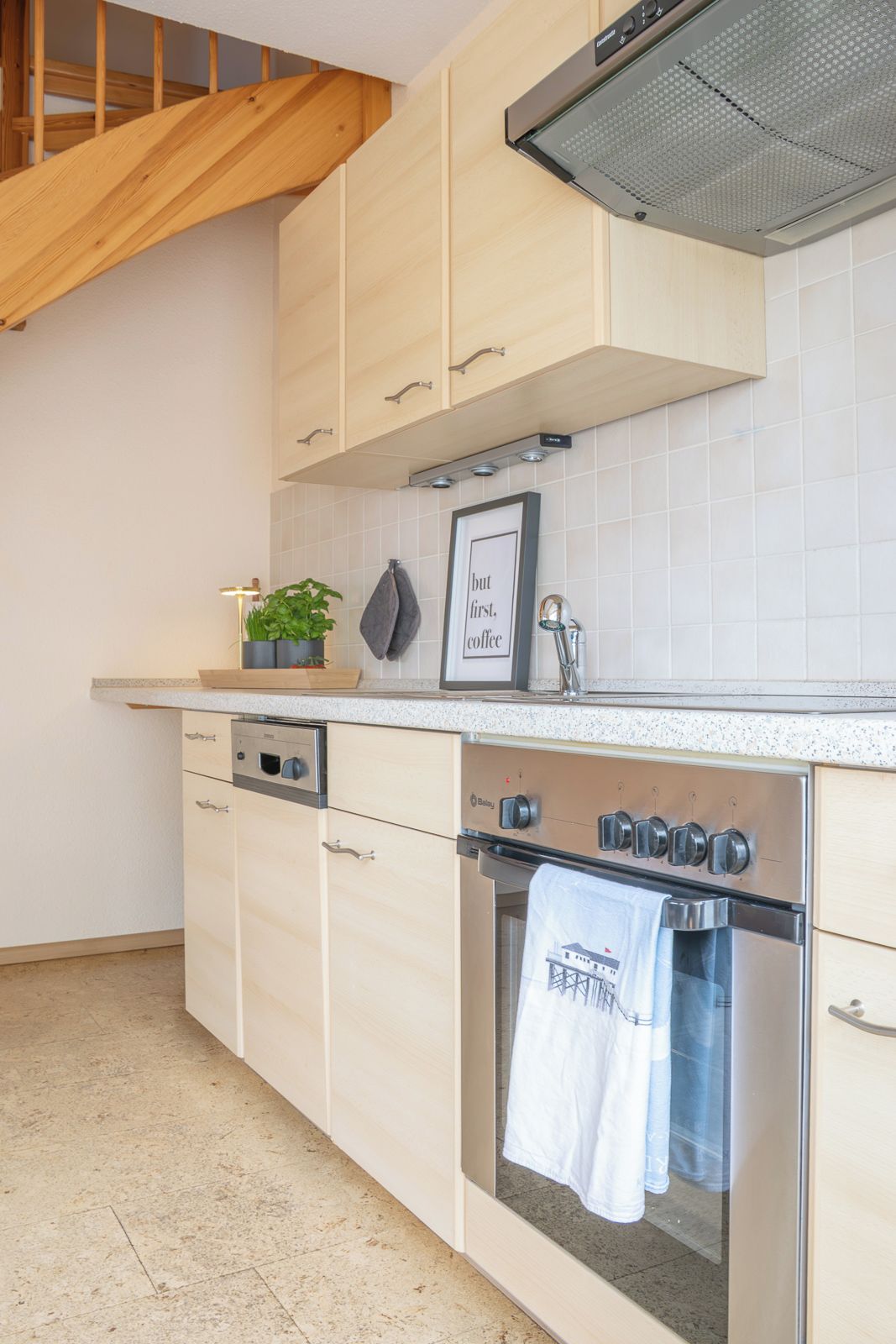 Een keuken met lichte houten keukenkastjes, een roestvrijstalen oven en een lijst met een citaat aan de muur.