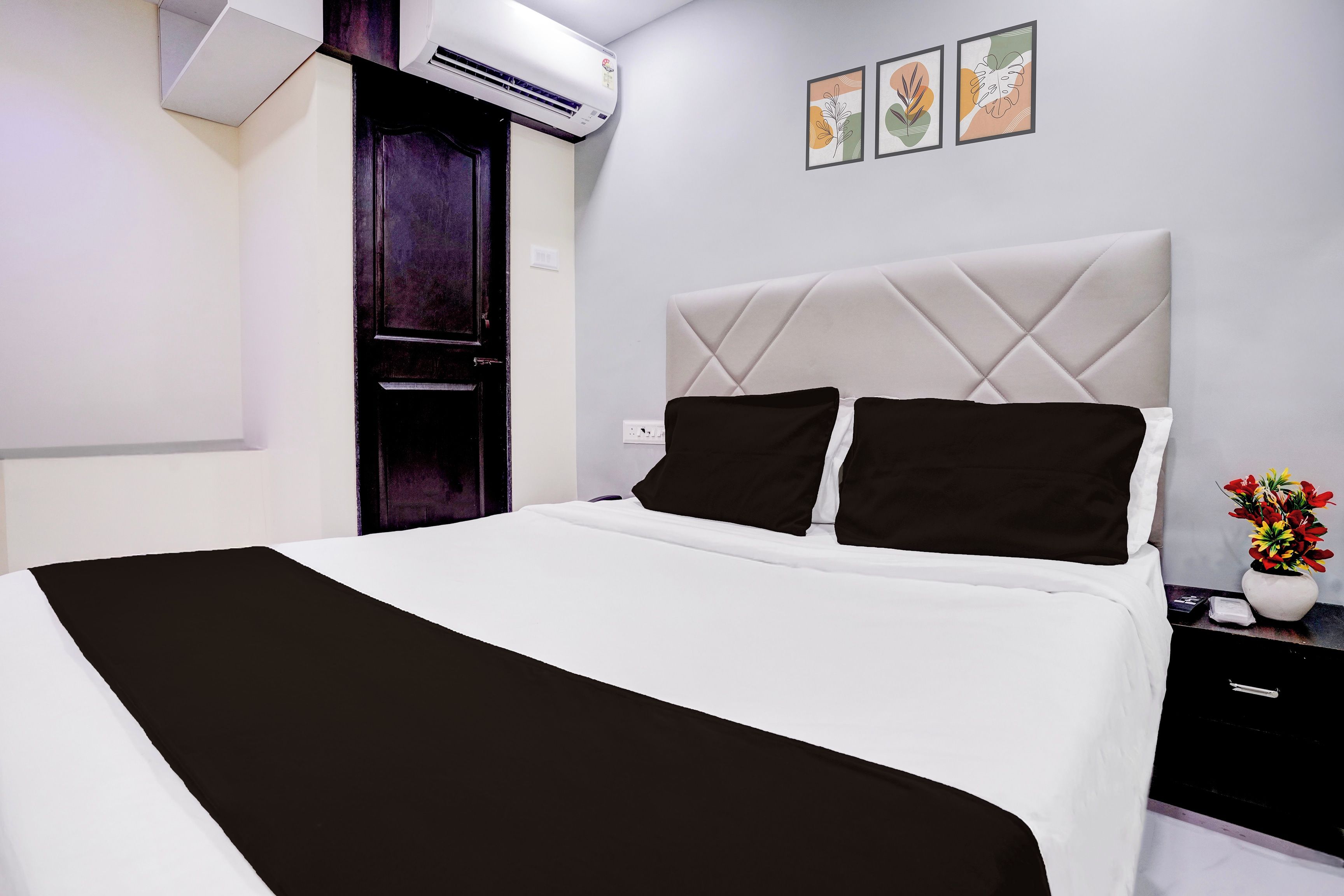 A hotel room with a bed, a nightstand, a vase with flowers, and a wall with three framed pictures.