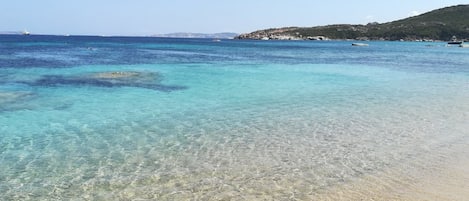 Una playa con aguas cristalinas y poco profundas, una orilla arenosa y una costa rocosa en la distancia.