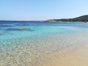 En strand med klart, lavt vand, sandet bredde og en fjerntliggende kystlinje af klippefyldte områder.