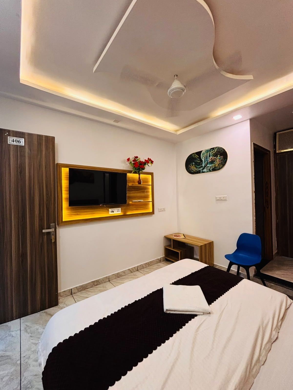 Habitación de hotel con una cama, televisor montado en la pared, un pequeño escritorio, una silla y una puerta con el símbolo del número.