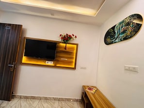 A room with a wall-mounted TV, a wooden bench, a small table with a magazine, and a painting on the wall. - Raj Rani Complex And Hotel Restaurant (Mainpuri)