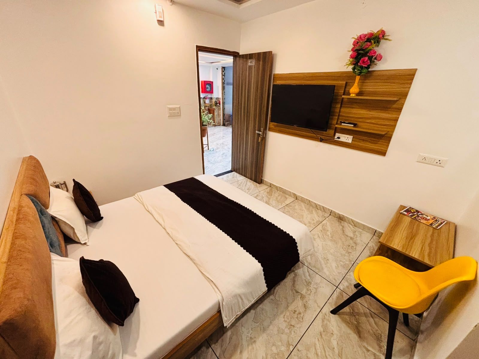 Habitación de hotel con una cama, un televisor montado en la pared, una mesita de noche de madera y una silla amarilla.