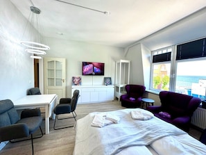 Ein modernes Hotelzimmer mit Bett, Schreibtisch, Stuhl, violettem Sessel, Fernseher und Meerblick.