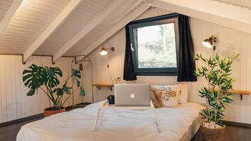 A bedroom with a sloped ceiling, a bed with a laptop, a window with curtains, and potted plants.