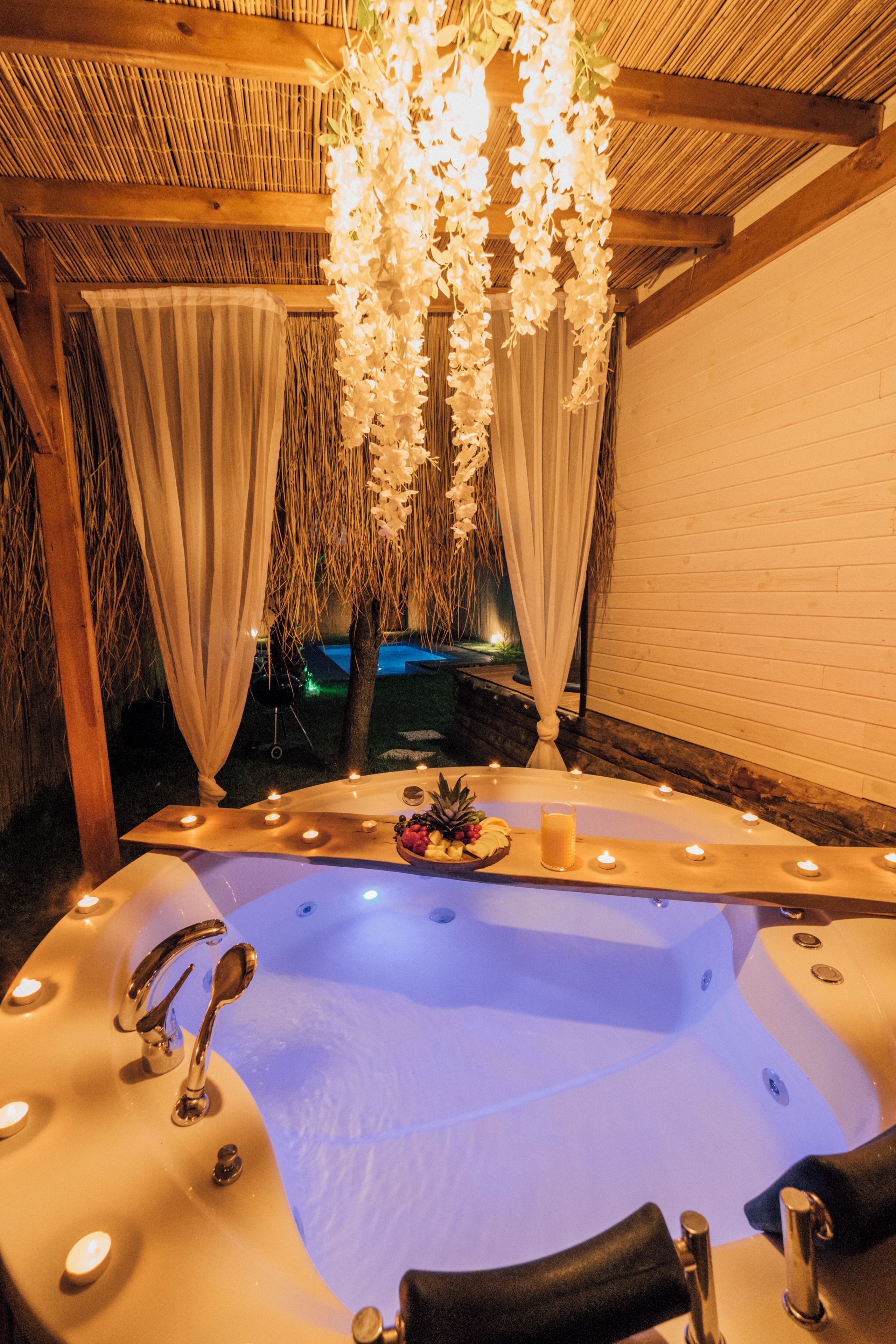 A hot tub with candles and fruits, surrounded by a wooden pergola and white curtains.