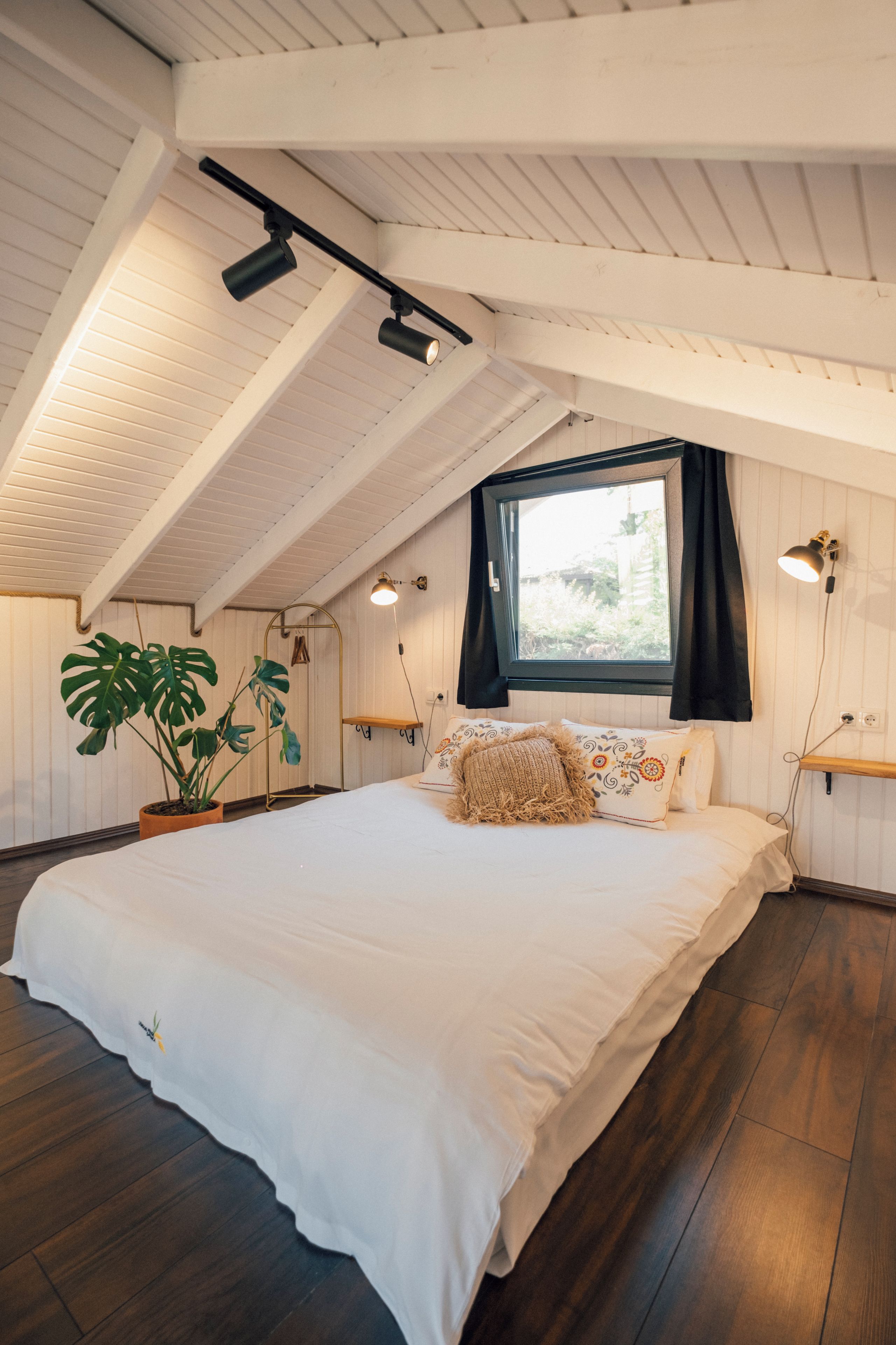 A bedroom with a large bed, a window with curtains, a potted plant, and a wooden ceiling.