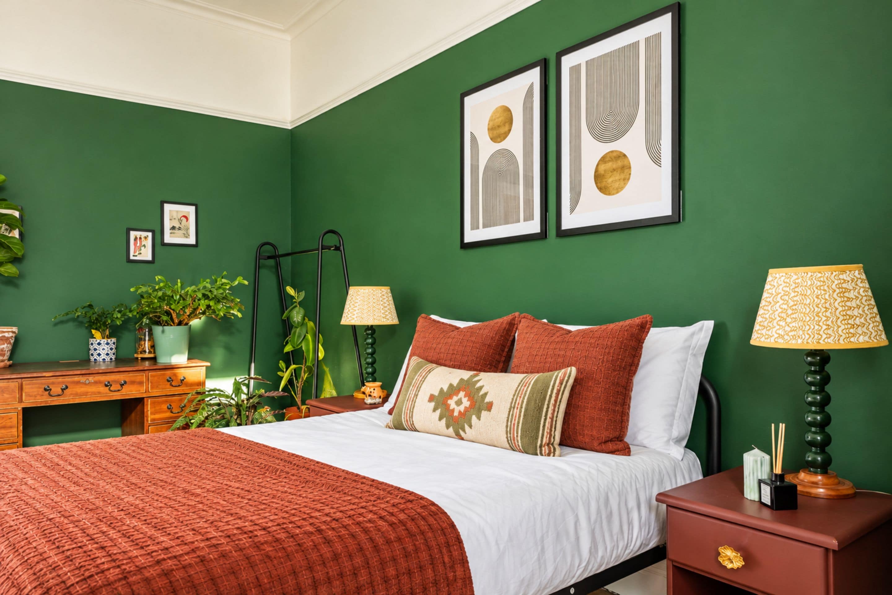 Een slaapkamer met een groene wand, een bed met veel kussens, een nachtkastje met een lamp en een dressoir met planten.