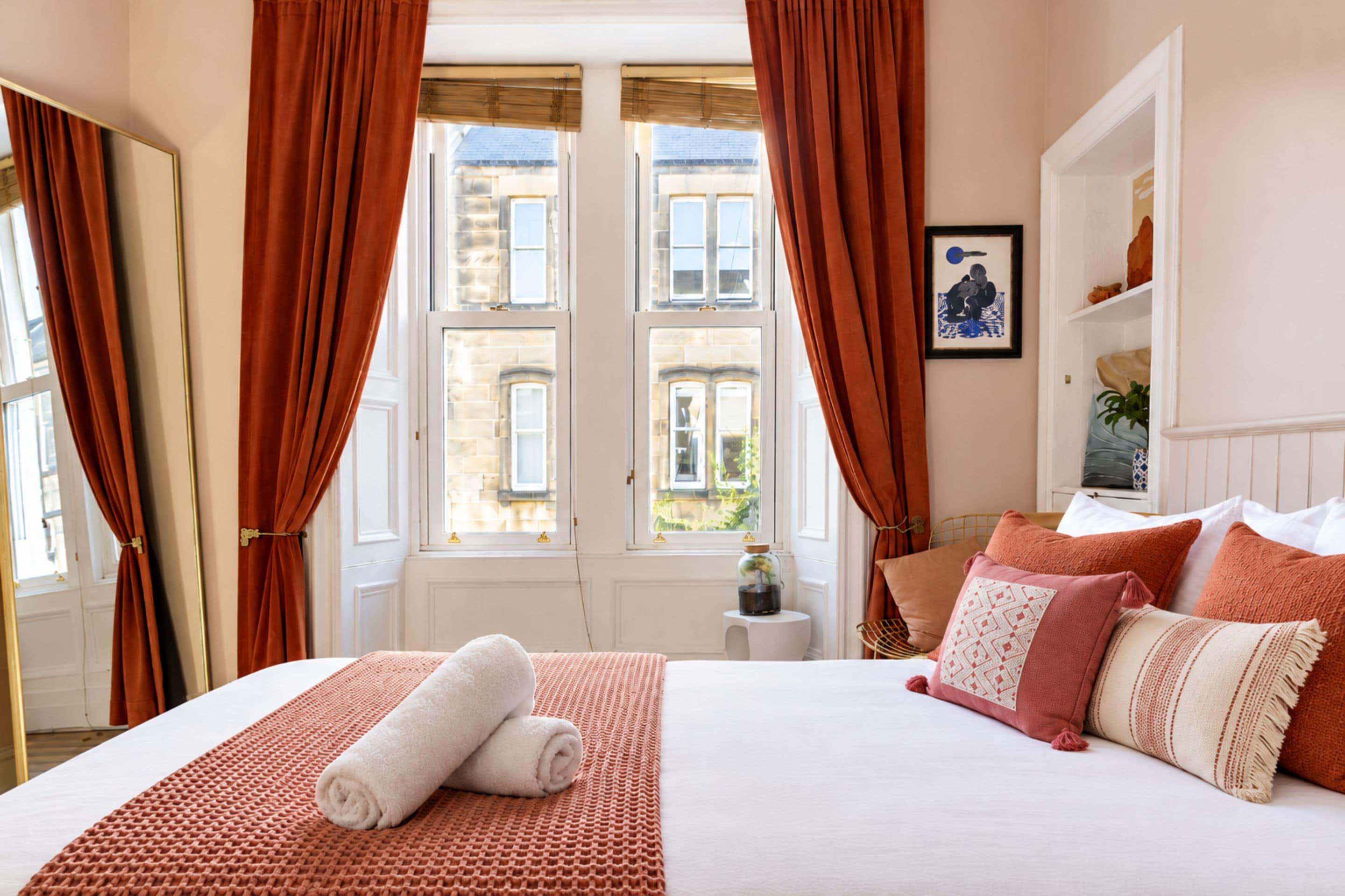 Quarto de hotel com cama, janela com cortinas, mesa pequena com um vaso e um quadro na parede.