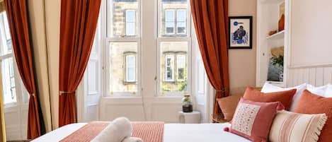 A hotel room with a bed, a window with curtains, a small table with a vase, and a wall with a framed picture.